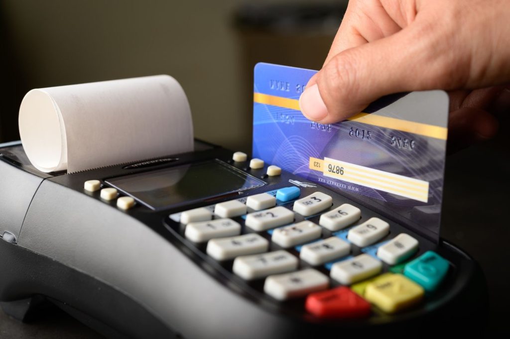A hand swipes a blue credit card through a point-of-sale terminal, a transaction that directly impacts the user's current credit utilization ratio.
