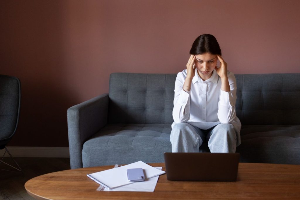An anxious woman sits on a couch holding her head while looking at a laptop after having her loan denied.