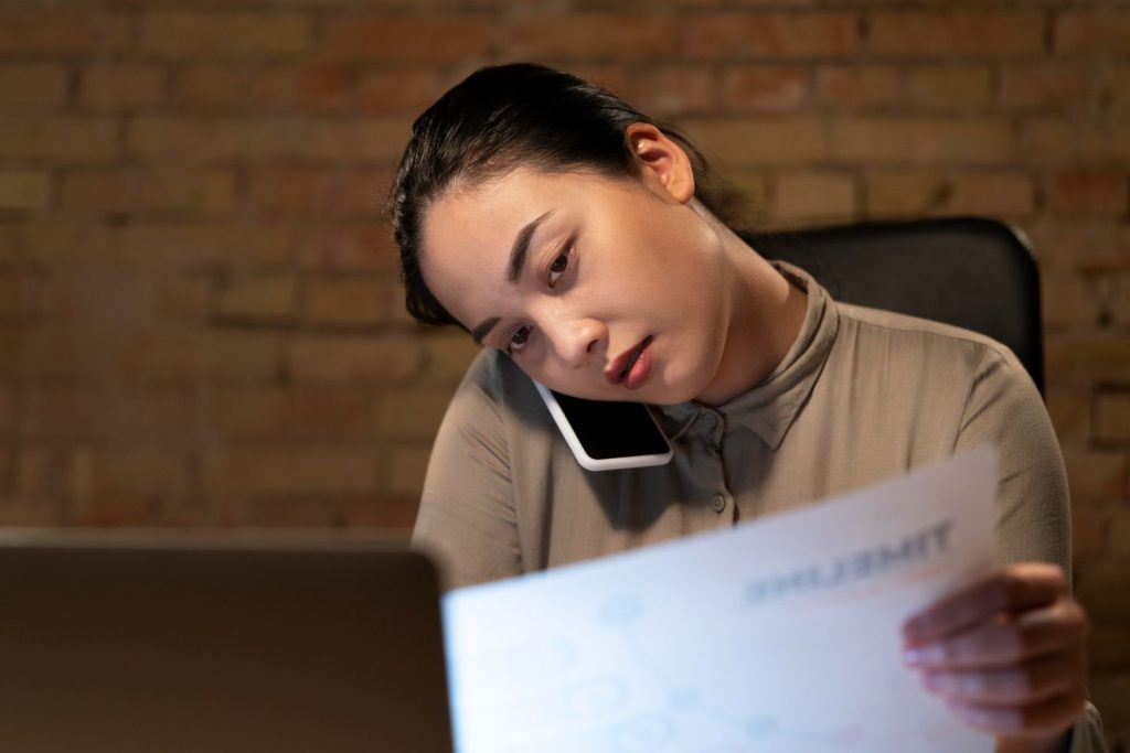 A tired woman reviews financial documents and talks on the phone late at night after a loan denied notification.
