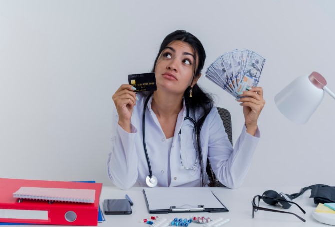 Thoughtful young female doctor wearing medical robe and stethoscope sitting at desk with medical tools holding money ands credit card analyzing her unnecessary expenses