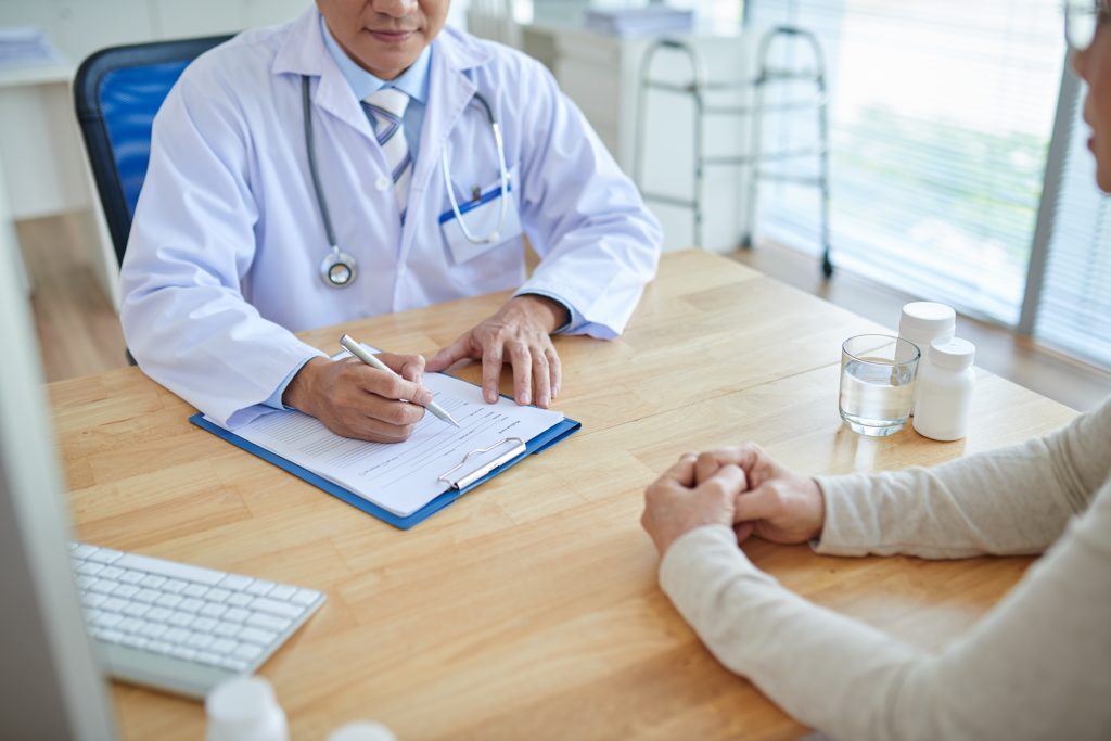 Close-up shot of male doctor filling in out-of-pocket maximum medical record, his patient sitting opposite him