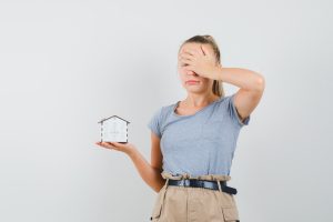 Young lady in t-shirt, pants holding house model and looking sorry , front view.