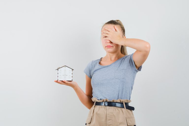 Young lady in t-shirt, pants holding house model and looking sorry , front view.