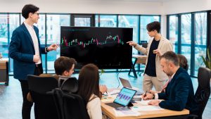 Two young workers leading business meeting in an office, discussing the topic of cryptocurrencies with other workers using a big display with currency rate, physical coins, and proprietary trading