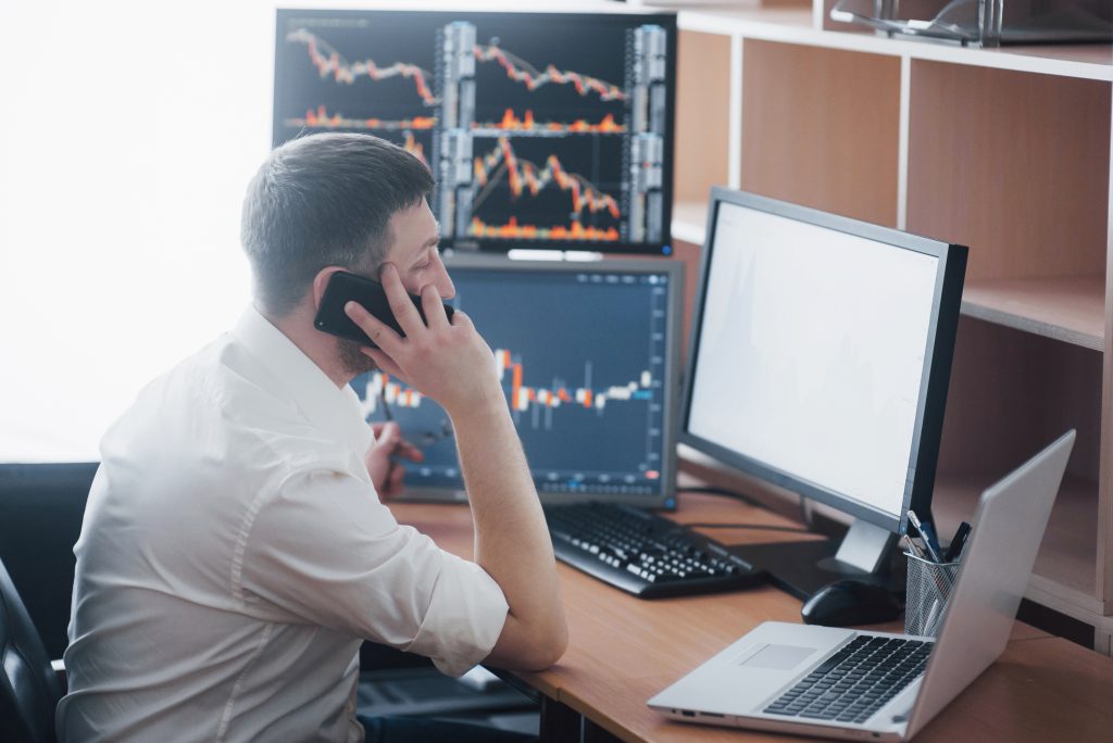 Over the shoulder view of and stock broker trading online while accepting orders by phone. Multiple computer screens ful of charts and data analyses in background.