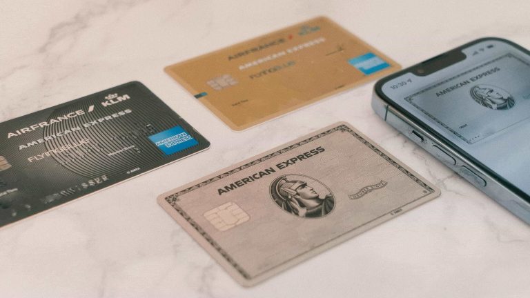 Multiple American Express credit cards, including Air France KLM Flying Blue and classic Amex designs, displayed on a marble surface next to a smartphone with a digital AmEx card