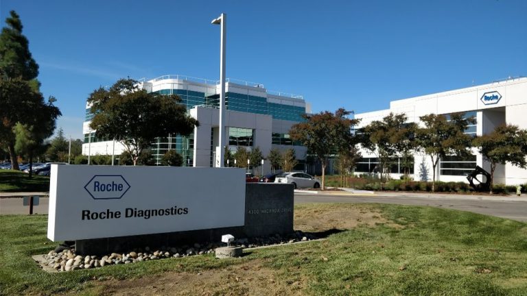 Roche Diagnostics headquarters building with company sign at 4300 Hacienda Drive, modern glass offices surrounded by trees.
