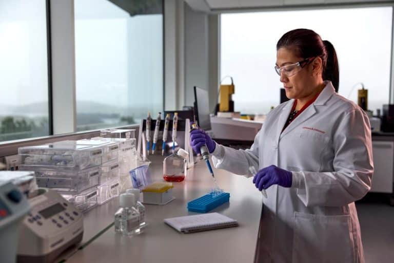 Scientist in a Johnson & Johnson lab using a pipette to prepare samples for biomedical research, wearing a lab coat, gloves, and safety glasses in a modern laboratory setting.