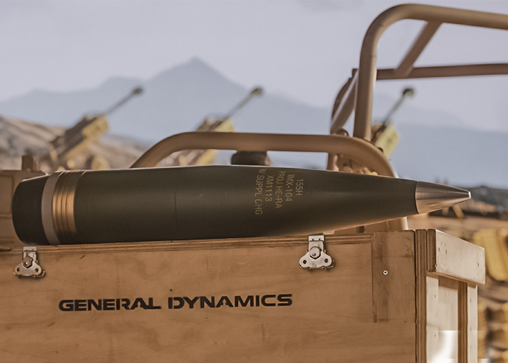 General Dynamics 155mm artillery shell on a wooden crate, with howitzers in a desert test range background.