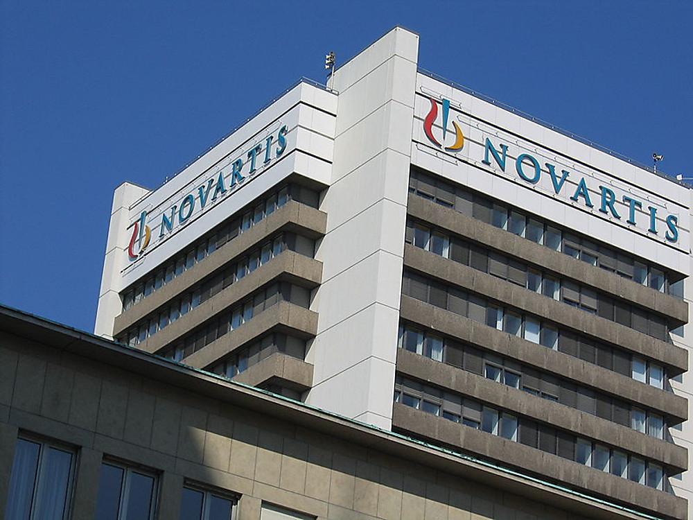 Novartis headquarters building in Basel, Switzerland, featuring the company logo on a modern glass and concrete facade under a clear blue sky.