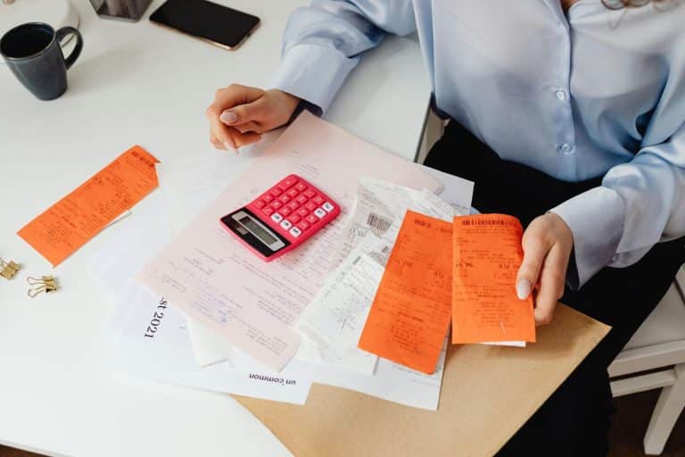 A person organizing bills, receipts, and rent documents with a calculator on a white desk while calculating monthly expenses and checking if utilities are included in rent.