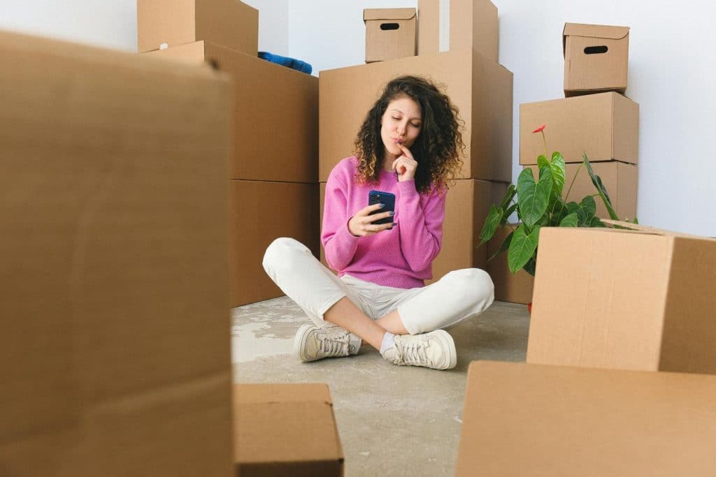New homeowner sitting on the floor surrounded by moving boxes, using smartphone while unpacking in her new home.