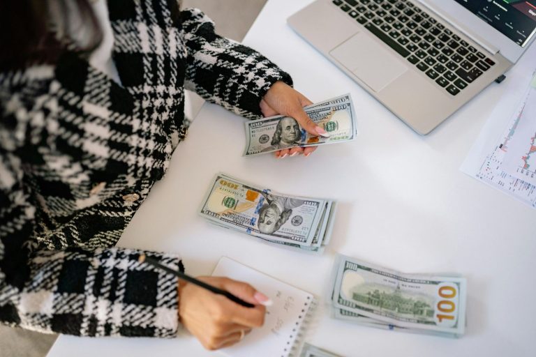 Person counting hundred-dollar bills at a desk with a laptop and notes while calculating savings for a rainy day fund.