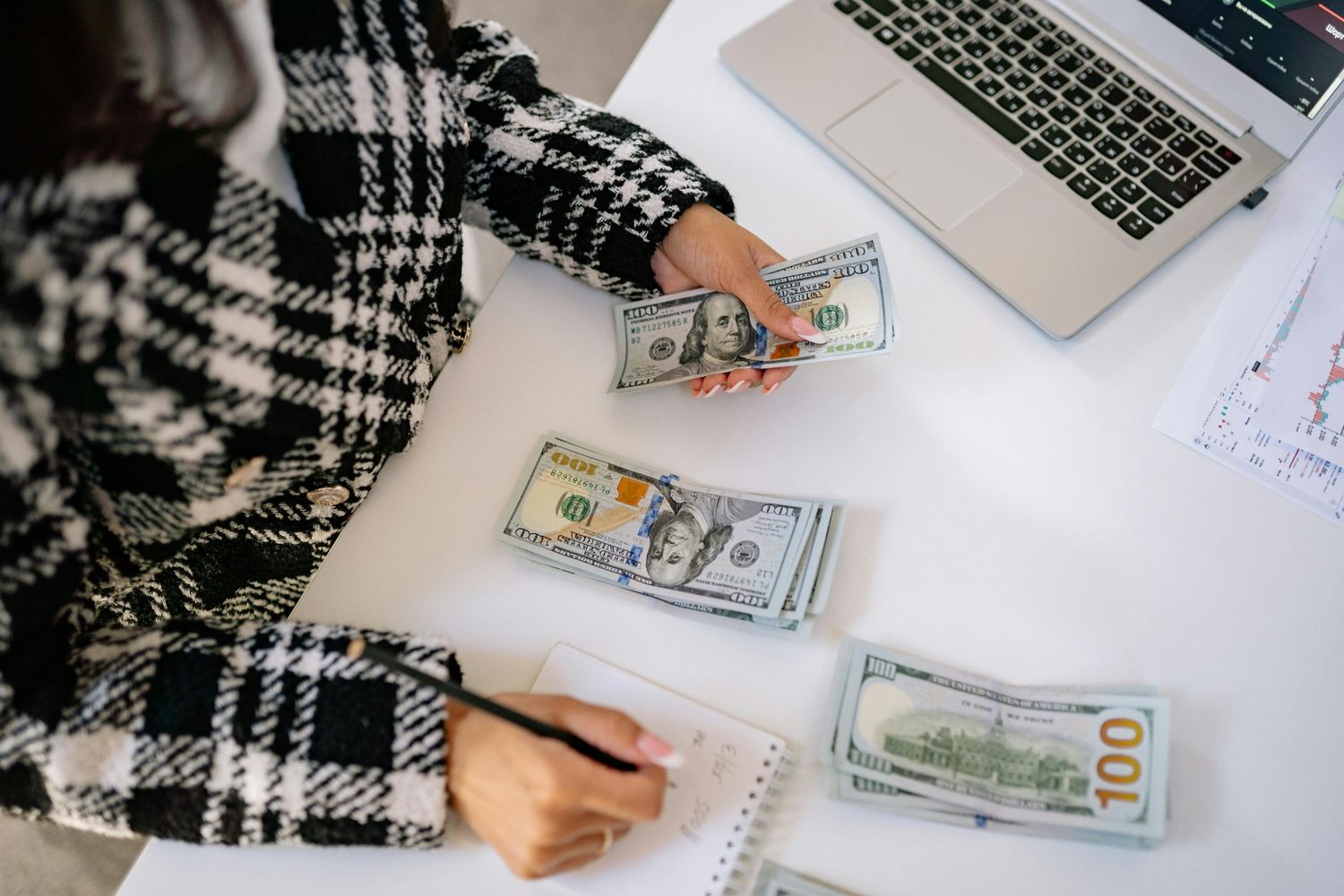 Person counting hundred-dollar bills at a desk with a laptop and notes while calculating savings for a rainy day fund.