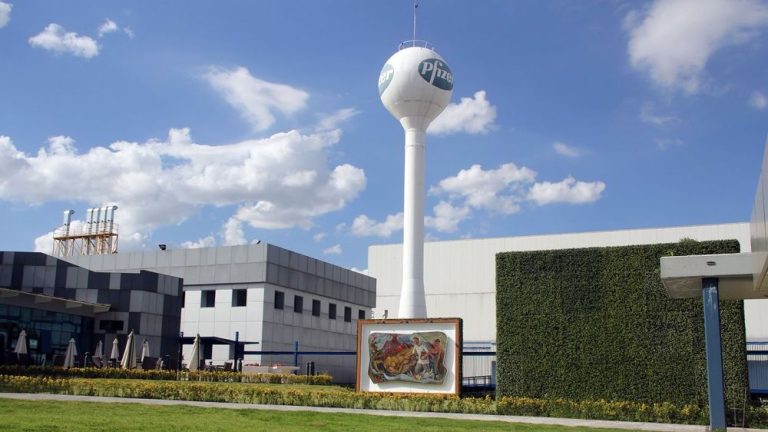 Exterior view of Pfizer manufacturing facility with modern buildings, landscaped garden, and water tower displaying Pfizer logo under a clear blue sky.