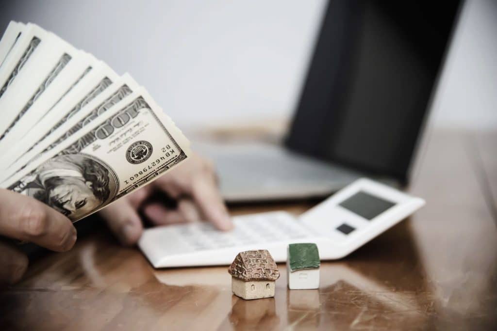 A person holding cash and using a calculator near small house models to budget for unsecured loans.