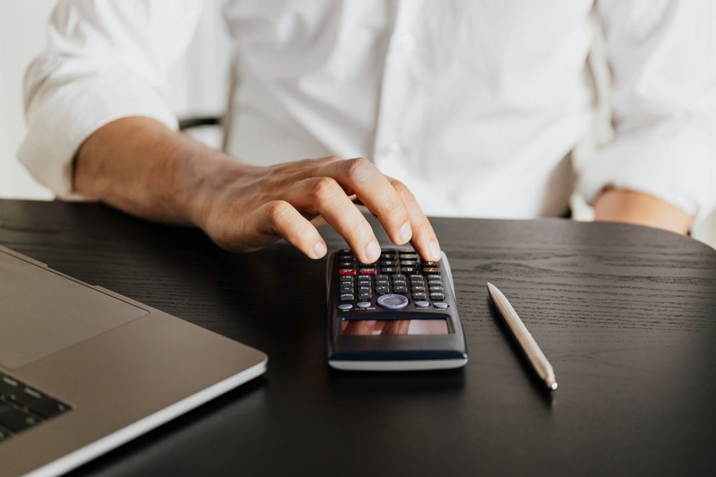 Close-up of a man using a calculator and laptop to determine the total cost of a loan origination fee.