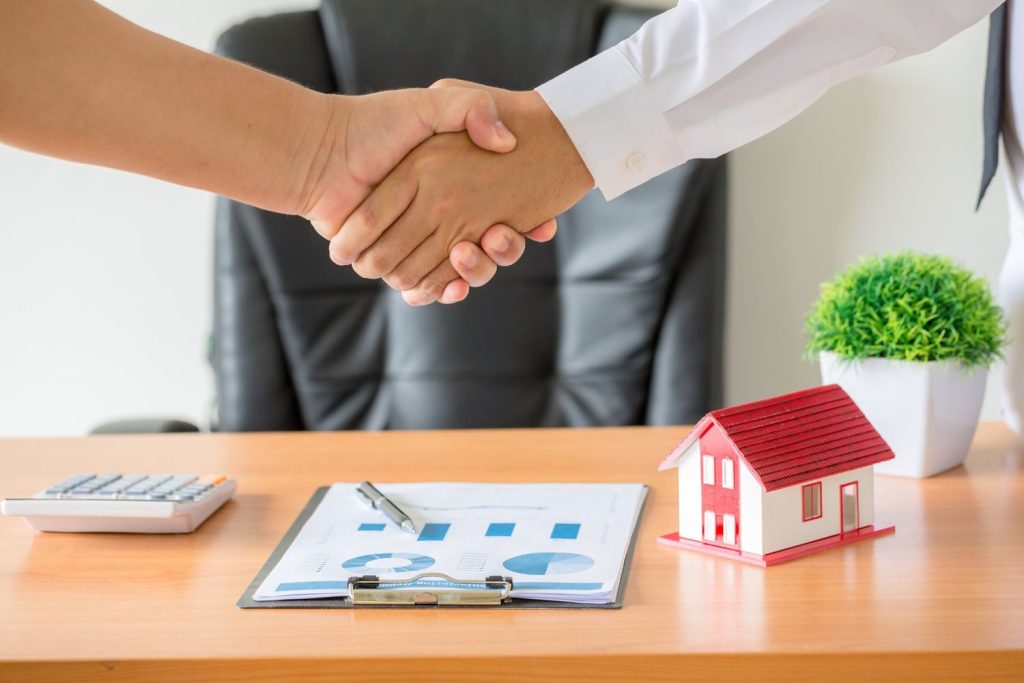 Two professionals shaking hands over a desk with a red-roofed house model and financial charts.