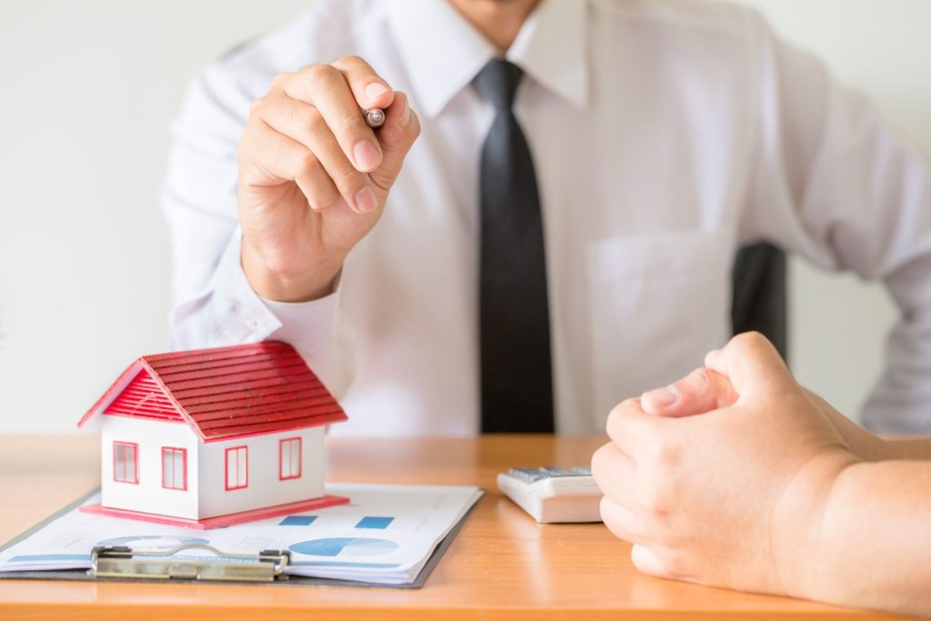 A real estate agent in a white shirt and tie pointing a pen toward a client to sign a mortgage agreement.
