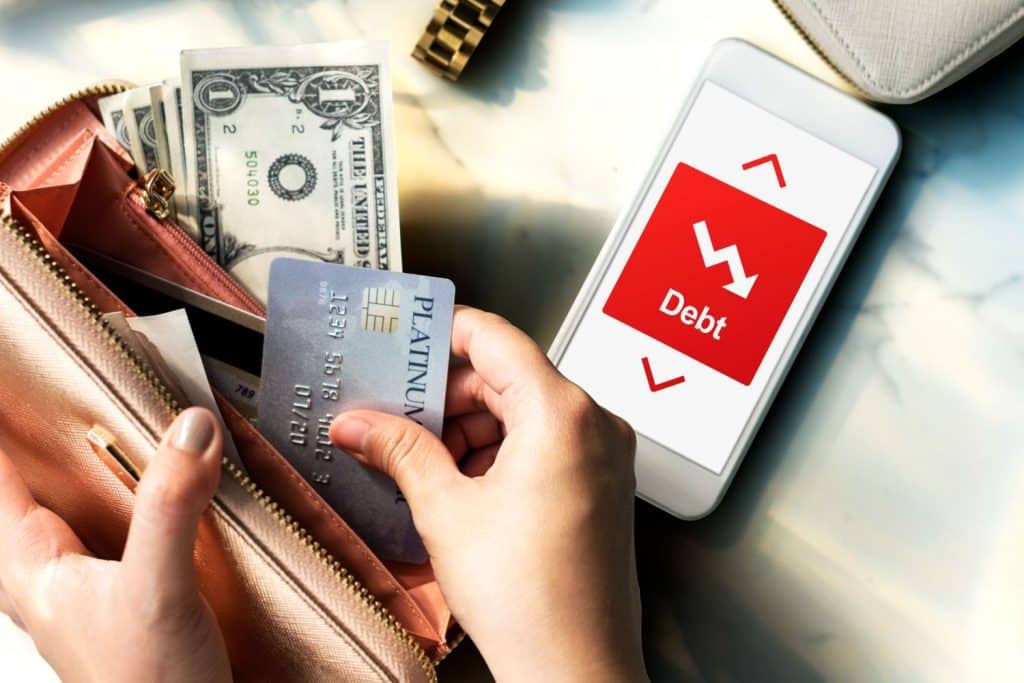 Woman holding credit card next to a phone displaying debt icons and cash in a wallet.