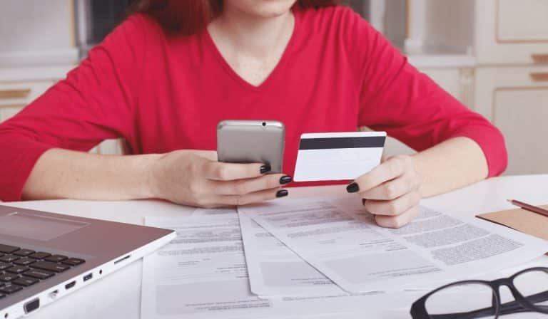 A woman holding a credit card and phone while managing paperwork.