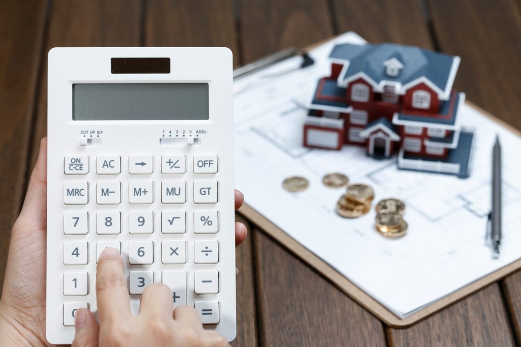 A person using a white calculator to figure out mortgage interest rates next to a villa house model and blueprints.