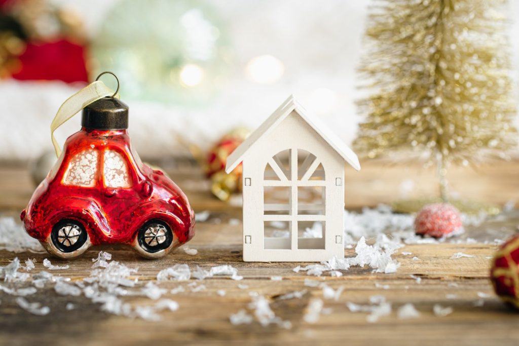 A red car ornament and a small white house on a snowy wooden surface, debating whether to buy a house or car.