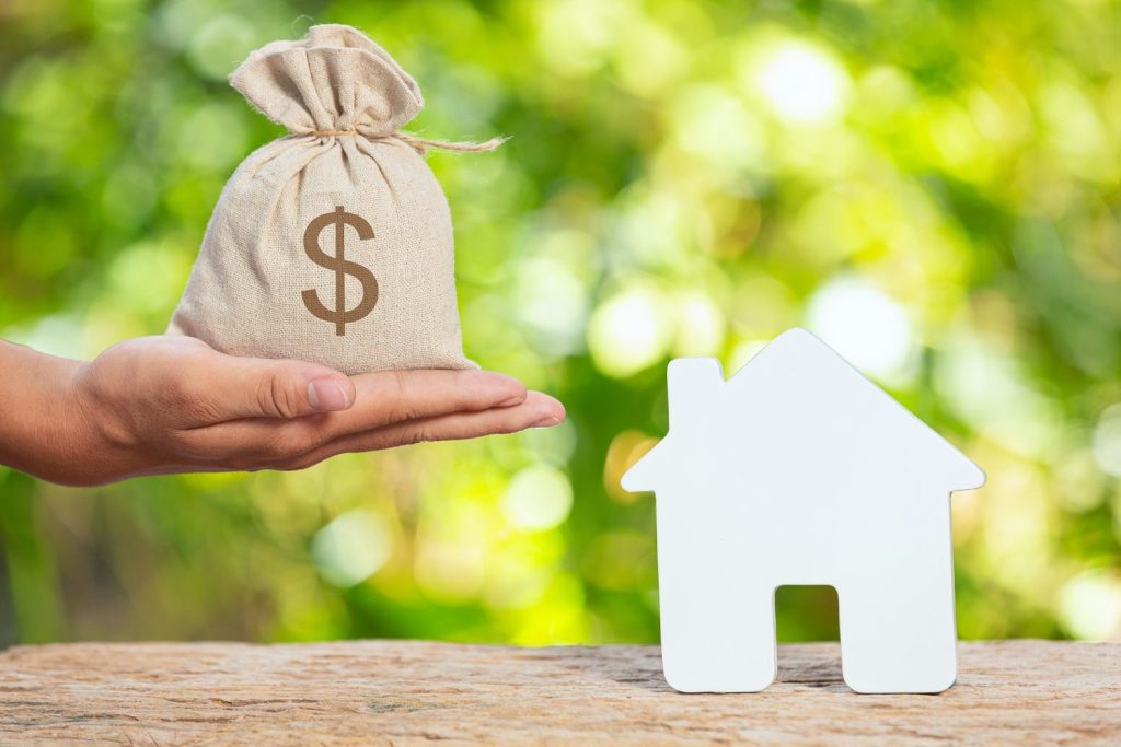 A hand holds a money bag next to a house cutout, representing liquidating equity through cash-out refinance.
