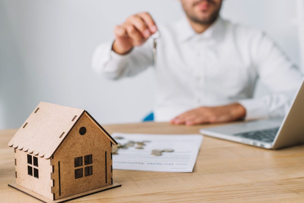 A mortgage broker holding up house keys next to a wooden model home and loan application documents.