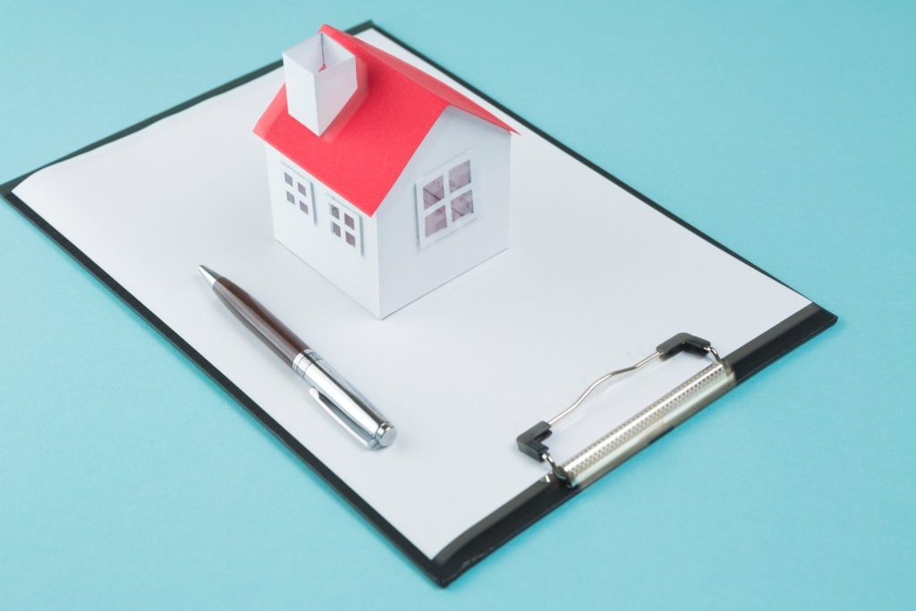 A paper house model sits on a blank clipboard, representing the initial preparation of a mortgage note agreement.