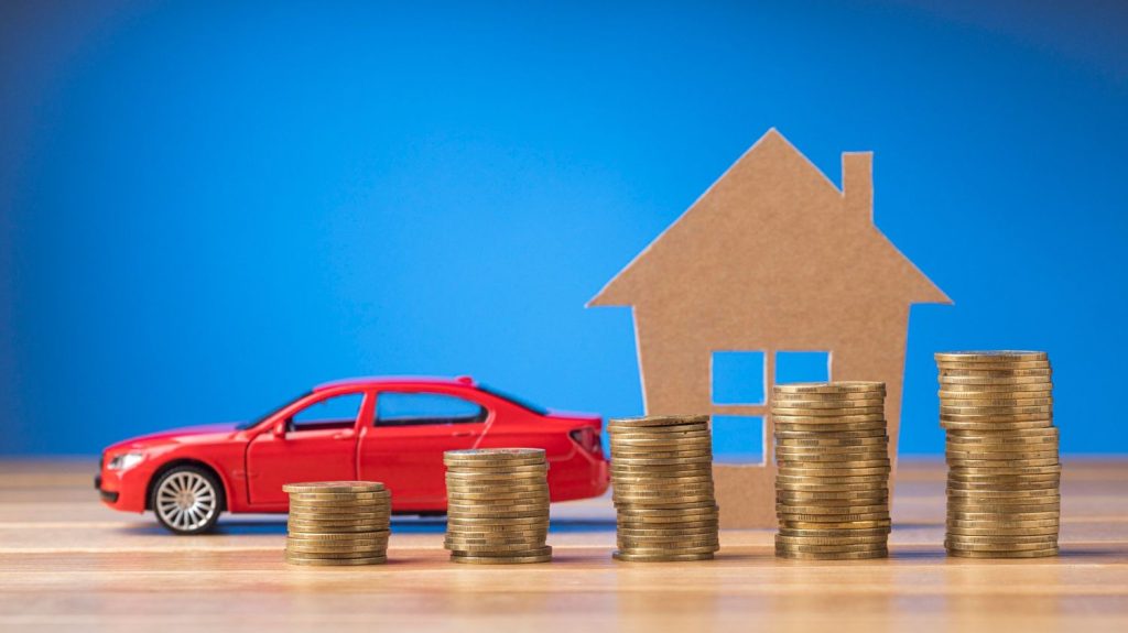 Piles of gold coins forming a graph in front of a red house or car cutout, symbolizing savings and investment.
