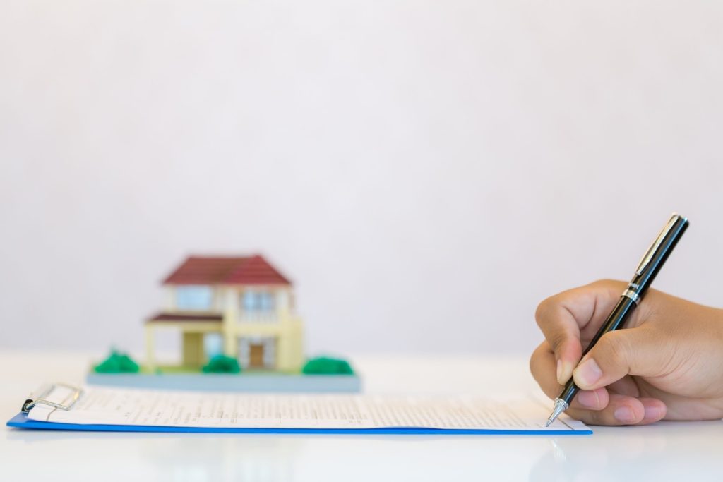 Close-up of a hand signing a mortgage note document with a blurred model house in the background.
