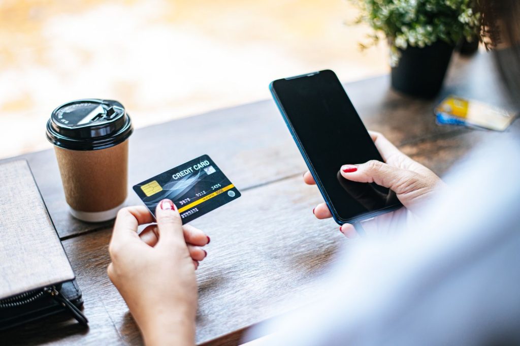 A person holding a credit card and smartphone at a table, representing how to use the best credit cards for groceries when shopping online or through delivery apps.