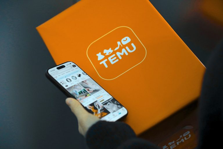 A person holding a smartphone open to the Temu app, with an orange Temu-branded package in the background.