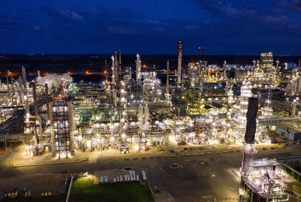 Brightly lit oil refinery complex at night with towers, pipelines, and processing units.