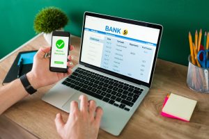 Person using a phone and laptop with an online banking dashboard and a “money received” screen.