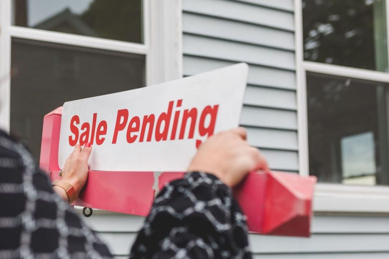 Person placing a real estate sign reading "Sale Pending" outside a house, illustrating pending home sales activity.