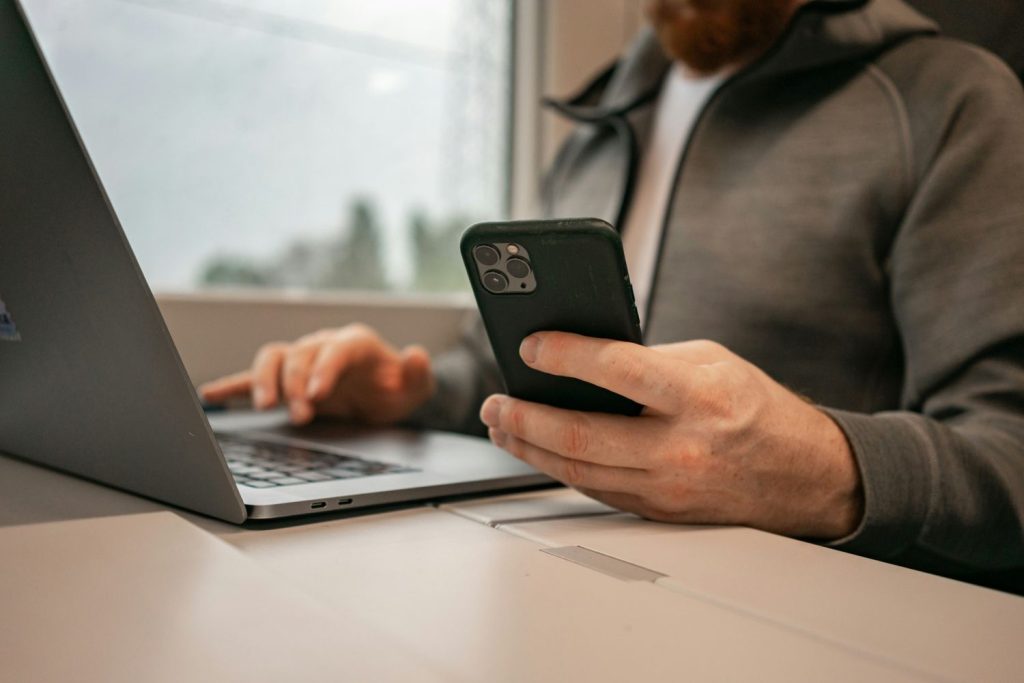 Person holding a smartphone while using a laptop at a desk checking their savings account.