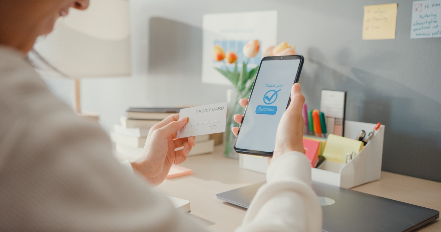 Person using a smartphone wallet app and credit card for a checking account purchase at a desk.
