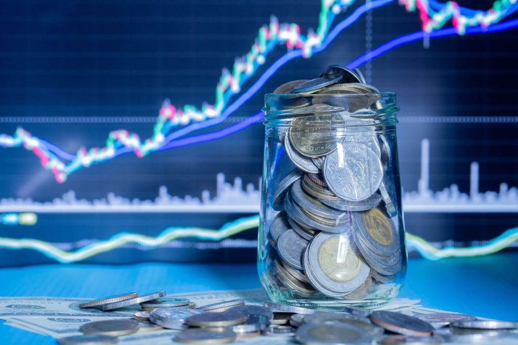 A jar filled with coins in front of stock charts represents saving regularly in an index fund.